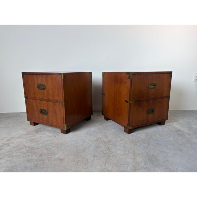 A handsome pair of vintage Baker Furniture campaign style nightstands featuring rich wood, recessed brass hardware, and...