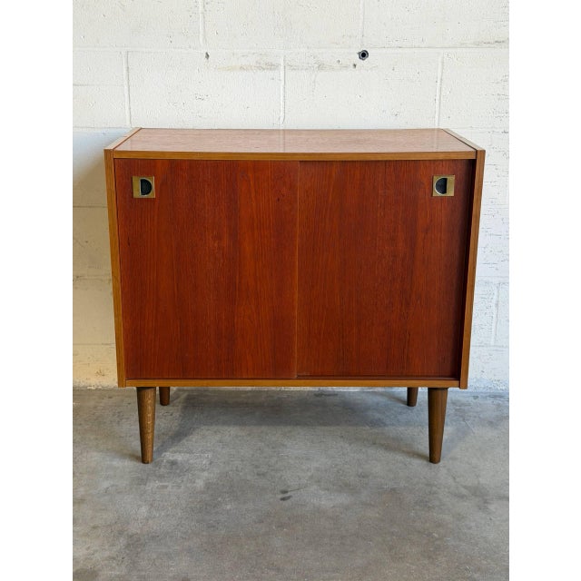 This compact storage piece features a clean rectilinear form with smooth sliding doors revealing interior shelving. Its...