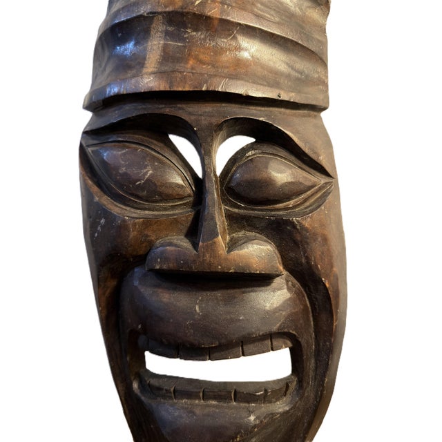Hand-carved wooden wall mask featuring an elongated face, tiered crescent headdress, and a fierce, grimacing expression...