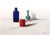 This artwork features a serene composition of three objects: a red spool of thread, a green vase, and a blue bottle, each...