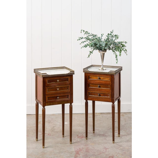 This elegant pair of Louis XVI style side tables features a brass gallery, three functional drawers, and lovely white/grey...