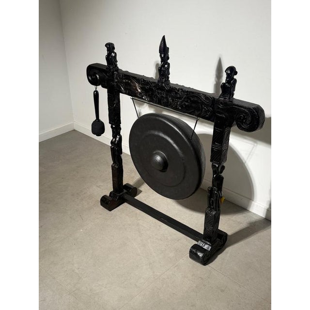 Striking freestanding metal gong suspended within an elaborately carved wooden frame. The dark-finished stand features...
