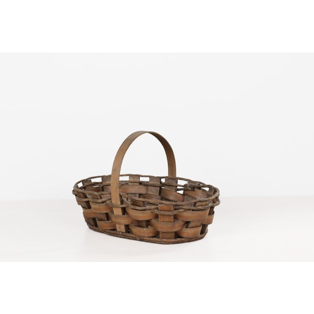 Vintage oval-shaped splint woven gathering basket featuring a solid wood base, and an arched wood handle. The sides are...