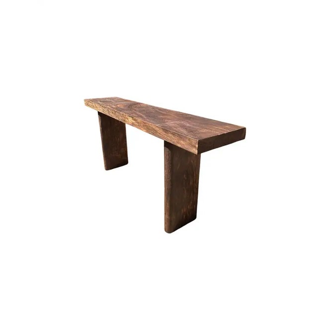 A sculptural mango wood console table crafted from a solid block of mango wood. The table top sits atop two solid, narrow...