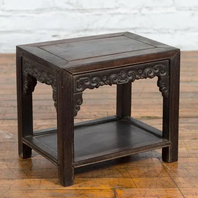 A Chinese Late Qing Dynasty period dark stool from the early 20th century with carved apron and lower shelf. Created in...