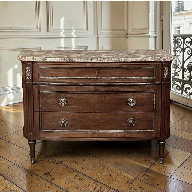 An antique French Louis XVI commode crafted from rich walnut and crowned with a luxuriously veined marble top in shades of...