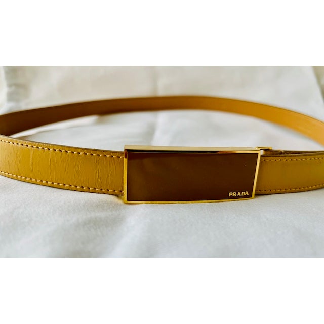 A vintage Prada leather belt in mustard/gold/tan, featuring a fully adjustable gold and brown enamel buckle stamped with...