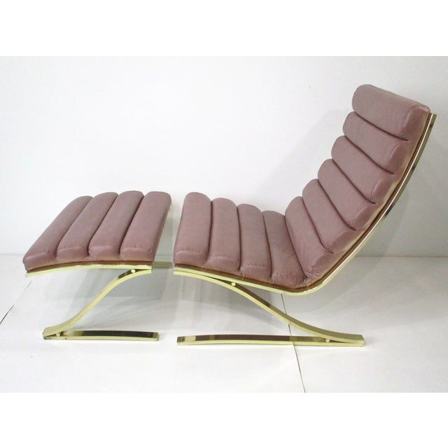 A two-piece vintage lounge chair with ottoman with heavy sculptural brass base and rolled leatherette upholstery in a gray...