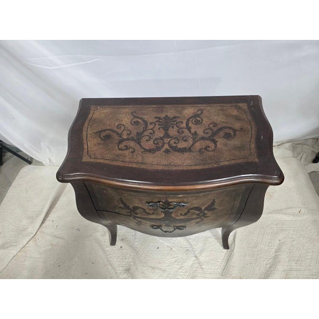 A vintage bombé-form two-drawer chest featuring a hand-painted decorative finish with scrolling motifs throughout. The...