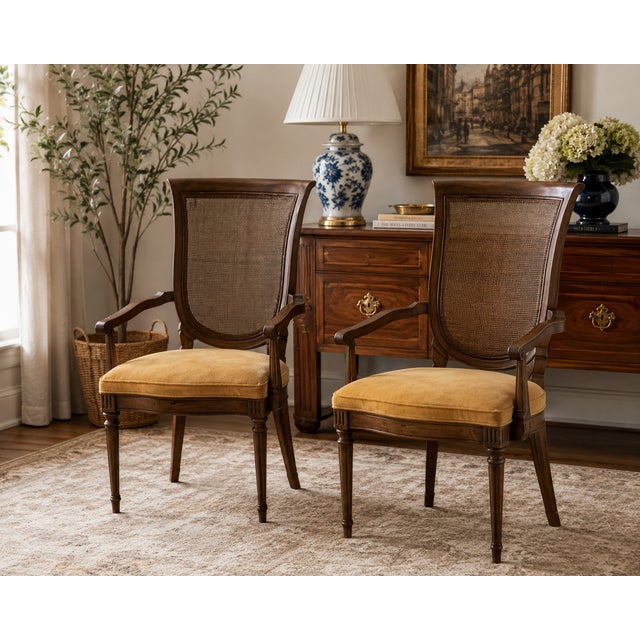 A sophisticated pair of vintage French Regency armchairs featuring elegant cane backs, sculpted walnut-toned frames, and...