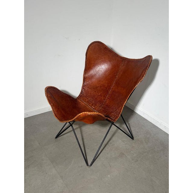 A distinctive butterfly lounge chair featuring a cognac brown leather sling seat on a slender black metal frame. Its...