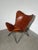 A distinctive butterfly lounge chair featuring a cognac brown leather sling seat on a slender black metal frame. Its...