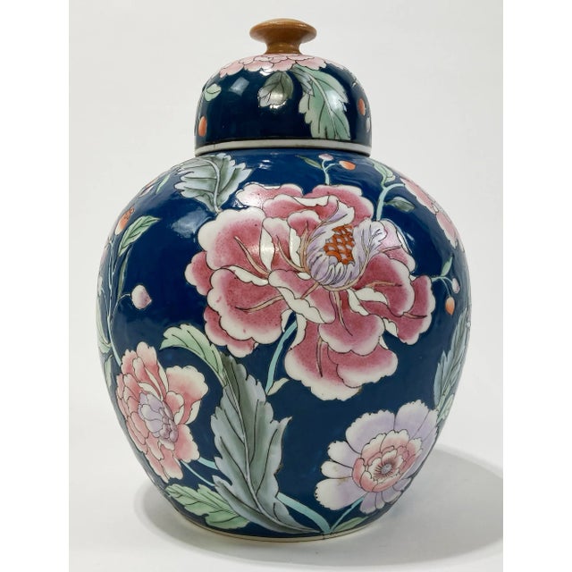 Deep blue ginger jar featuring large pink blossoms throughout body and lid. Lid has a wooden handle for easy removal....