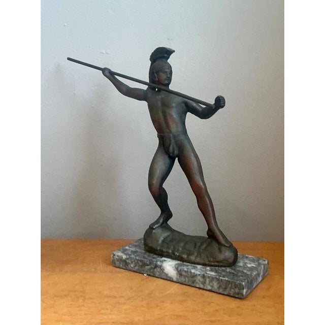 This vintage sculptural metal figure depicts a classical warrior in a dynamic, mid-action pose, one arm extended forward...