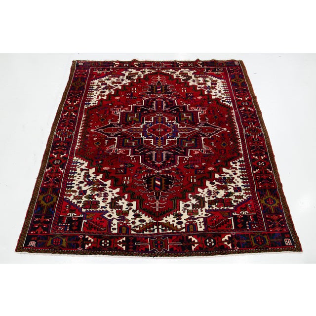This vintage Persian Heriz hand-knotted wool rug showcases a striking red field with bold blue and beige accents in an...