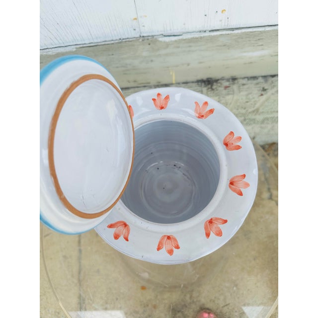 A small, hand-painted ceramic jar with a lid, featuring a soft pastel palette. The body has light blue vertical stripes...