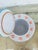 A small, hand-painted ceramic jar with a lid, featuring a soft pastel palette. The body has light blue vertical stripes...