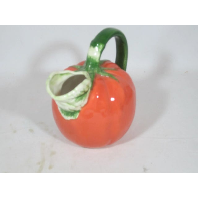 A vintage ceramic tomato pitcher, hand painted in a vibrant red with a green handle in the form of the tomato's stem and a...