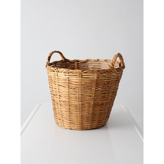 This vintage woven basket features a tall tapered form with double wrapped handles and a braided rim detail. The warm...
