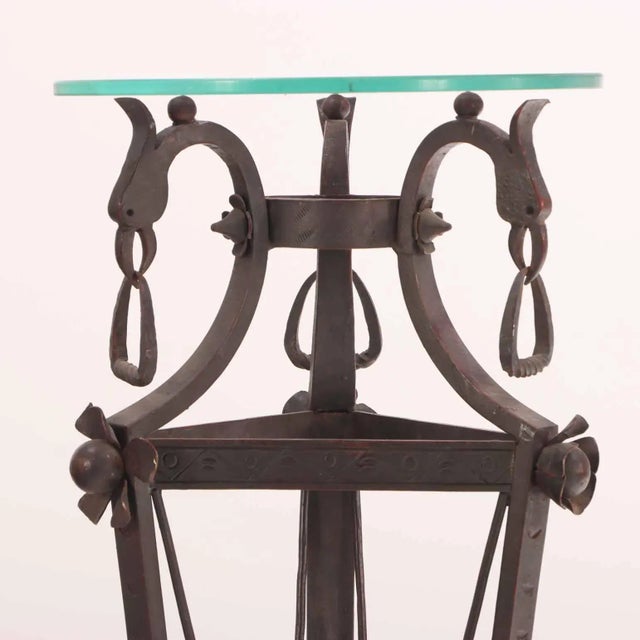 An elegant wrought iron plant stand or pedestal, featuring a round glass top supported by a tapering tripod base. The...