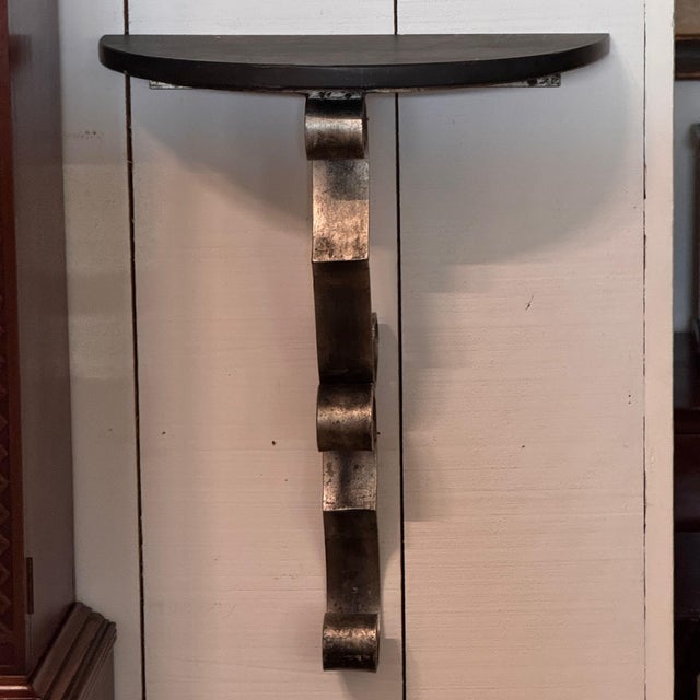 A wall mounted console with a demilune black marble top carried on a single steel bracket worked into a ribbon of deep...