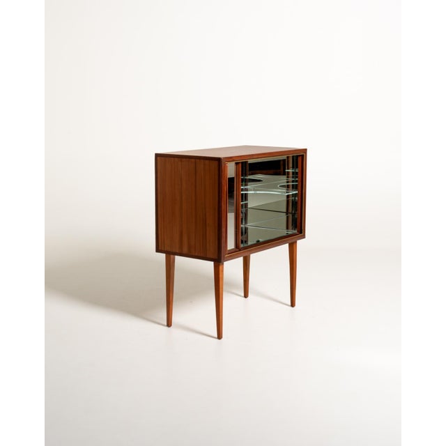 Scandinavian teak bar cabinet from the 1950s, with a sliding shutter door, brass trim, mirrored interior, and glass...