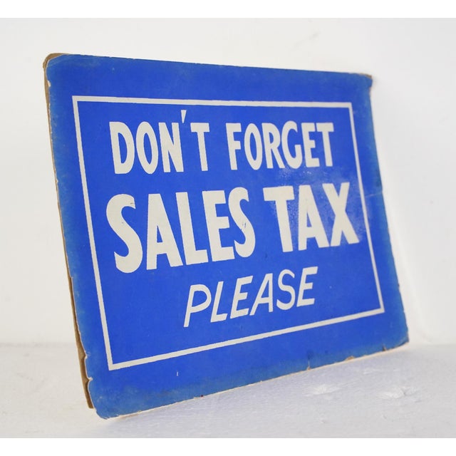 Vintage retail cardboard wall sign with a blue background and white lettering. The sign reads, Don't forget Sales Tax...