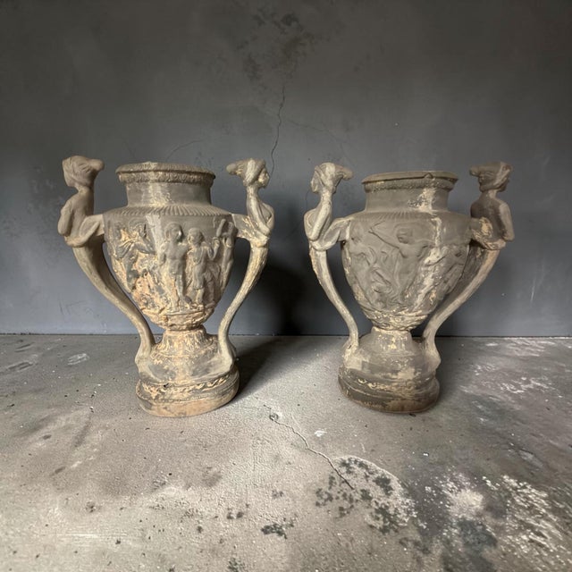 Striking pair of vintage neoclassical composition urns featuring detailed classical figures in relief and sculptural...