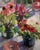 This artwork features a vibrant arrangement of potted flowers, including echinacea and other colorful blooms, set against...