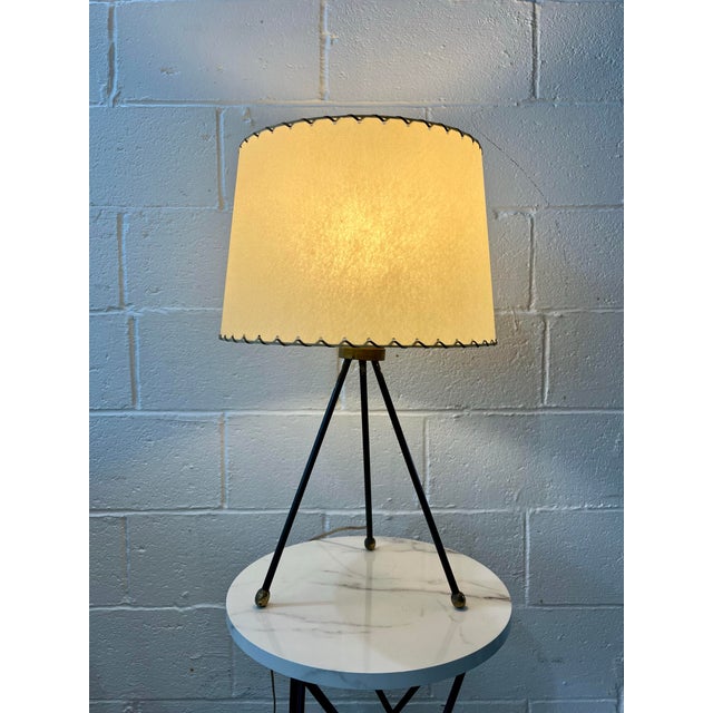 A sculptural mid-century modern tripod table lamp, featuring a slender blackened iron base accented with warm brass...