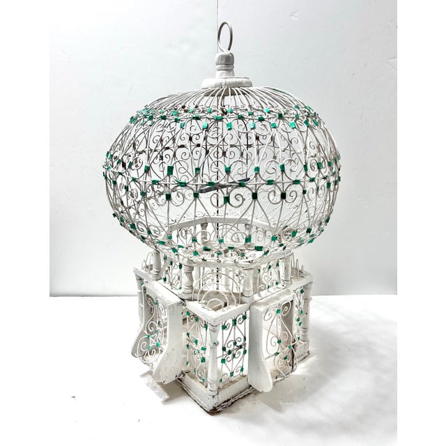 Ornate Hollywood Regency birdcage sculpture featuring intricate whitewashed metal scrollwork accented with green jewel...
