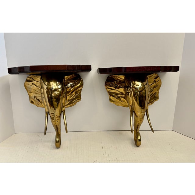 Pair vintage brass elephant head wall brackets featuring polished gold-tone faces and protruding tusks mounted into dark...