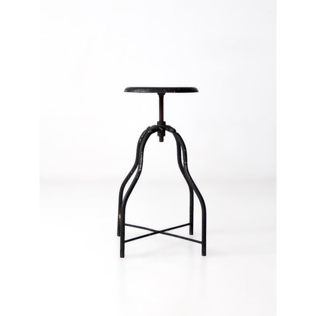 Early 20th-century adjustable industrial metal stool with a round screw-lift seat, curved steel rod legs, and simple cross...