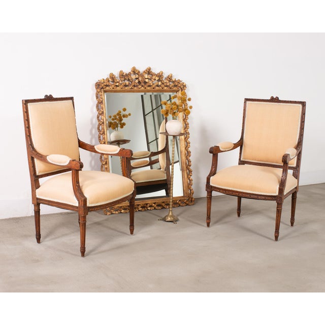 An elegant matched pair of walnut fauteuil armchairs in the grand Louis XVI style featuring classical hardwood frames...