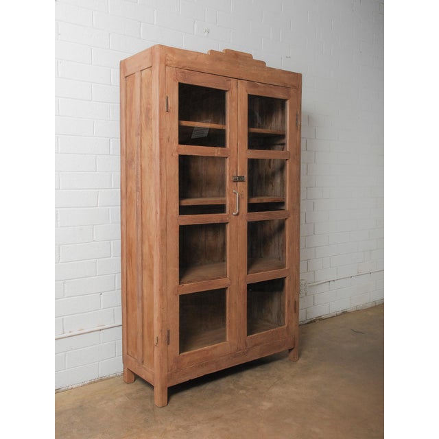 This substantial wooden glass-front almirah features a clean, rectilinear form softened by hand-worked surfaces and subtle...