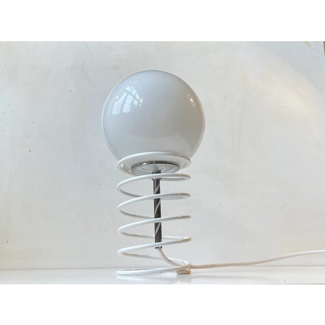 A simple small table light made from a steel spring in white lacquer freely holding an opaline glass sphere in a chrome...