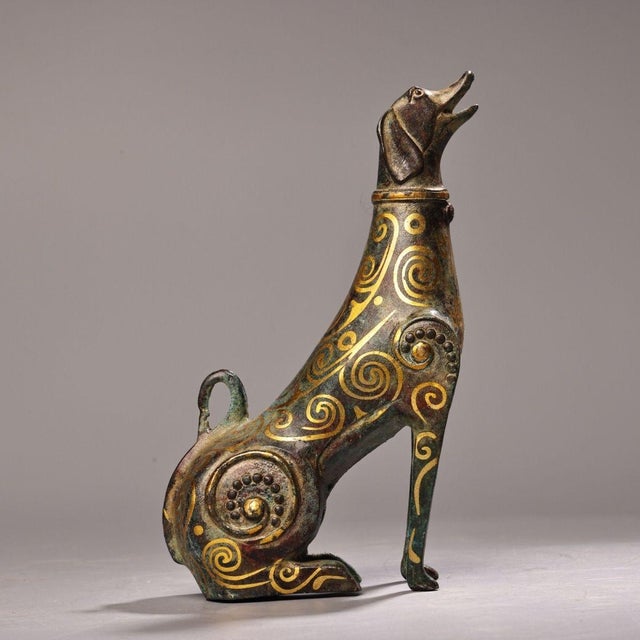 This is a cast bronze figurine of a seated dog, featuring extensive gold inlay (gilt bronze) decoration. The dog is...