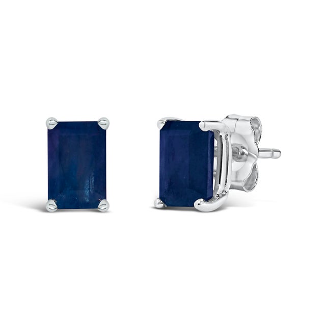Immerse yourself in the timeless elegance of these stud earrings, each boasting a lustrous 6x4mm emerald-cut blue...