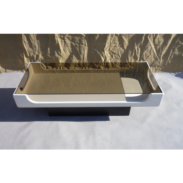 A Space Age style coffee table featuring a cabinet mounted on a base, topped with a rectangular smoked glass surface....