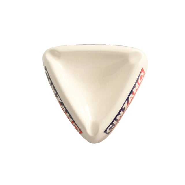 A triangular ceramic ashtray with the distinctive Cinzano logo in red, white and blue on all three sides, and a cigarette...