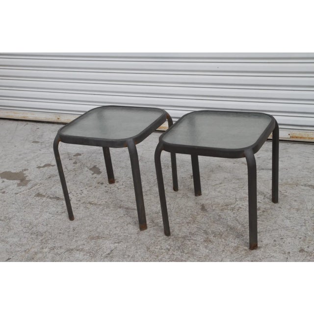 This is an outdoor end table with a metal frame and a glass top. The frame is black or dark gray with a tubular design,...