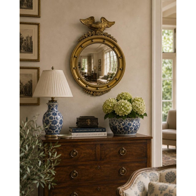 A Federal-style giltwood convex mirror crowned with a carved eagle, its wings outstretched above a laurel motif—an...