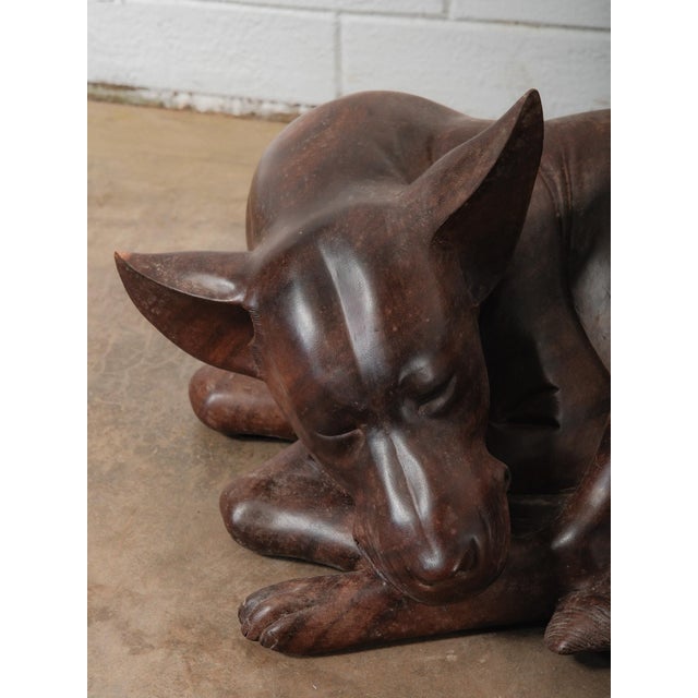 A striking hand-carved wooden sculpture depicting a resting dog, captured in a peaceful, curled pose. It's shown with its...