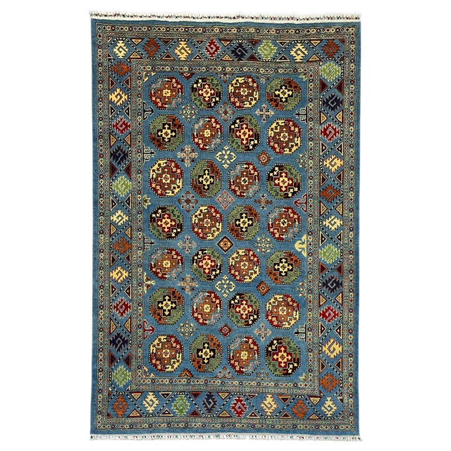 A blue wool area rug with a Bokhara-inspired tribal design. Rows of colorful octagonal gul motifs fill the blue field,...