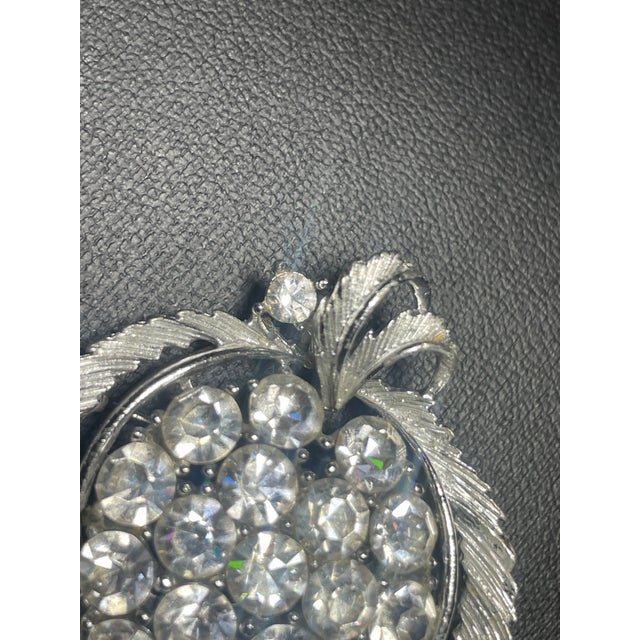 The brooch features a stylized leaf or heart outline framed by feathered, textured silver-tone leaves that curve upward...