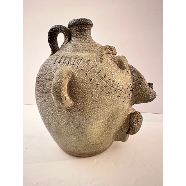 A face emerges from the body of this stoneware jug, built up in heavy relief rather than painted on. The speckled grey...
