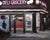 This oil painting captures a nighttime scene outside a deli grocery, illuminated by the bright neon lights that contrast...