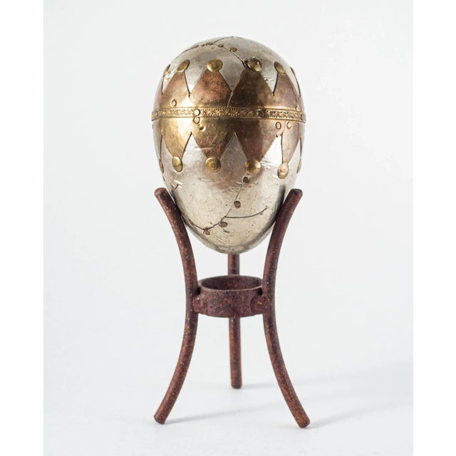 A sculptural bi-metal wrapped egg on tripod stand. Egg has copper and silver metal wrapped details, sections of metal...