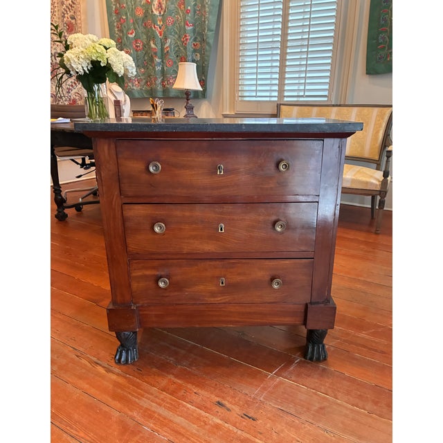 A French Empire three-drawer commode, circa 1810–1830, with tailored rectangular form and paw-foot base. Crafted from...