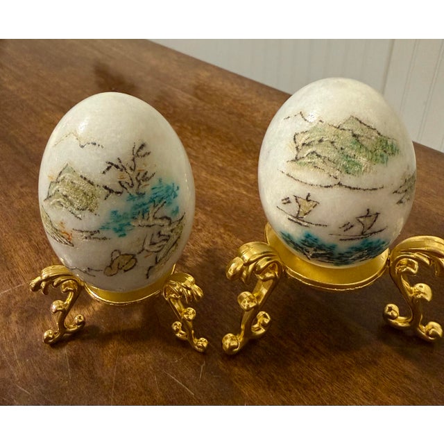 This pair of vintage decorative eggs showcases delicate hand-painted artistry, each resting on an ornate gold tone metal...